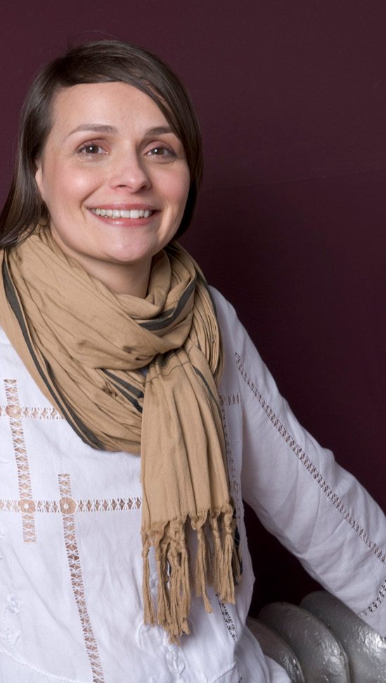 Alice Ritter smiling in a white embroidered top and a long camel scarf against a dark red wall