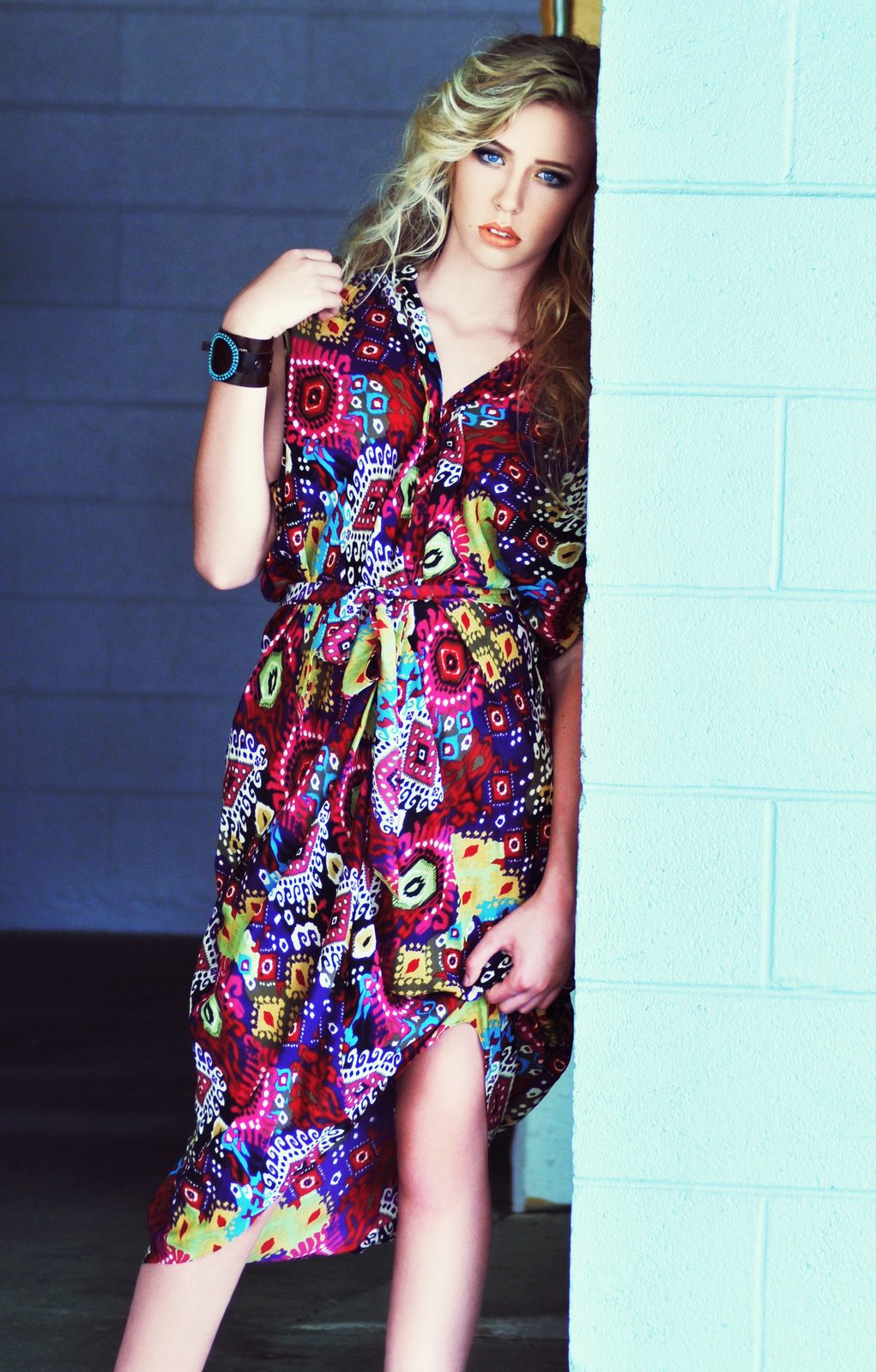 Model leaning against a mint green wall in a knee-length wrap dress with a bold multicolor graphic print