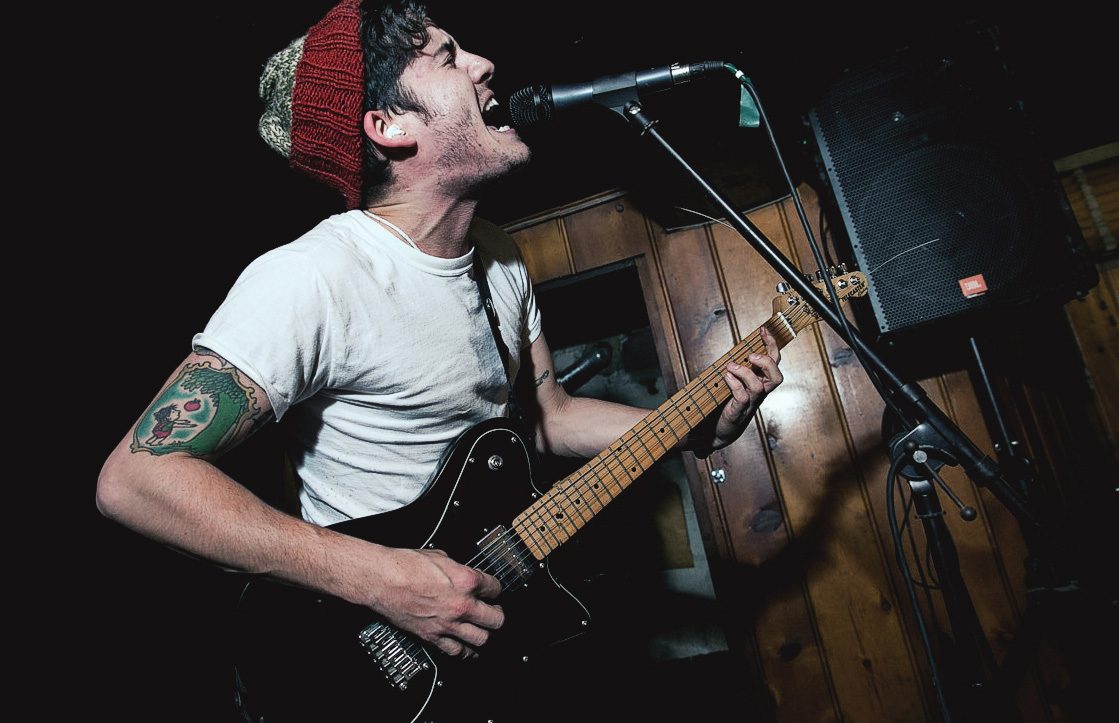 Collapser’s singer in a red knit beanie and white tee shouting into a mic while playing a black electric guitar in a wood-paneled room