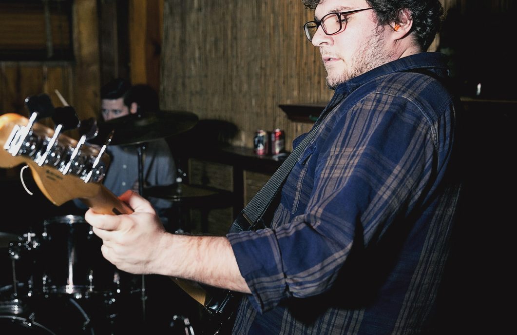 Collapser’s bassist in glasses and a plaid flannel shirt playing in front of the drummer in a wood-paneled basement