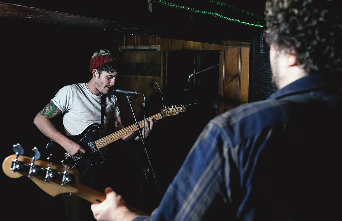 Collapser’s singer in a red beanie playing guitar at the mic, seen past the shoulder of the bassist in a plaid shirt