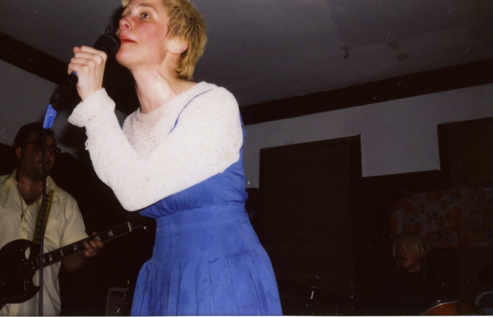 Priests’ singer with cropped blonde hair, a blue pinafore dress and white sweater singing into a handheld mic, a guitarist behind her