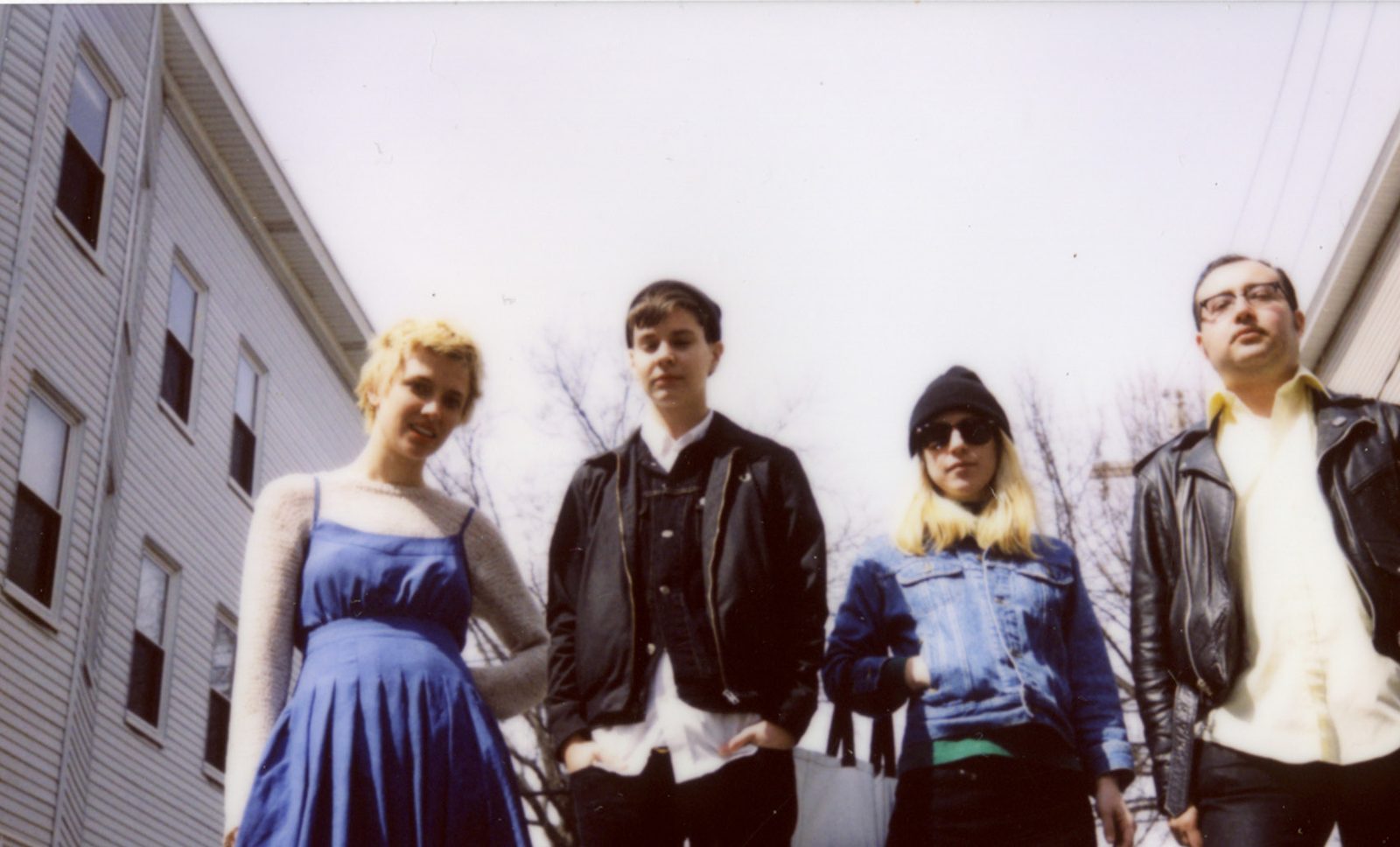 The four members of Priests in leather jackets, denim and a blue dress, posing on a street lined with clapboard houses under a white sky