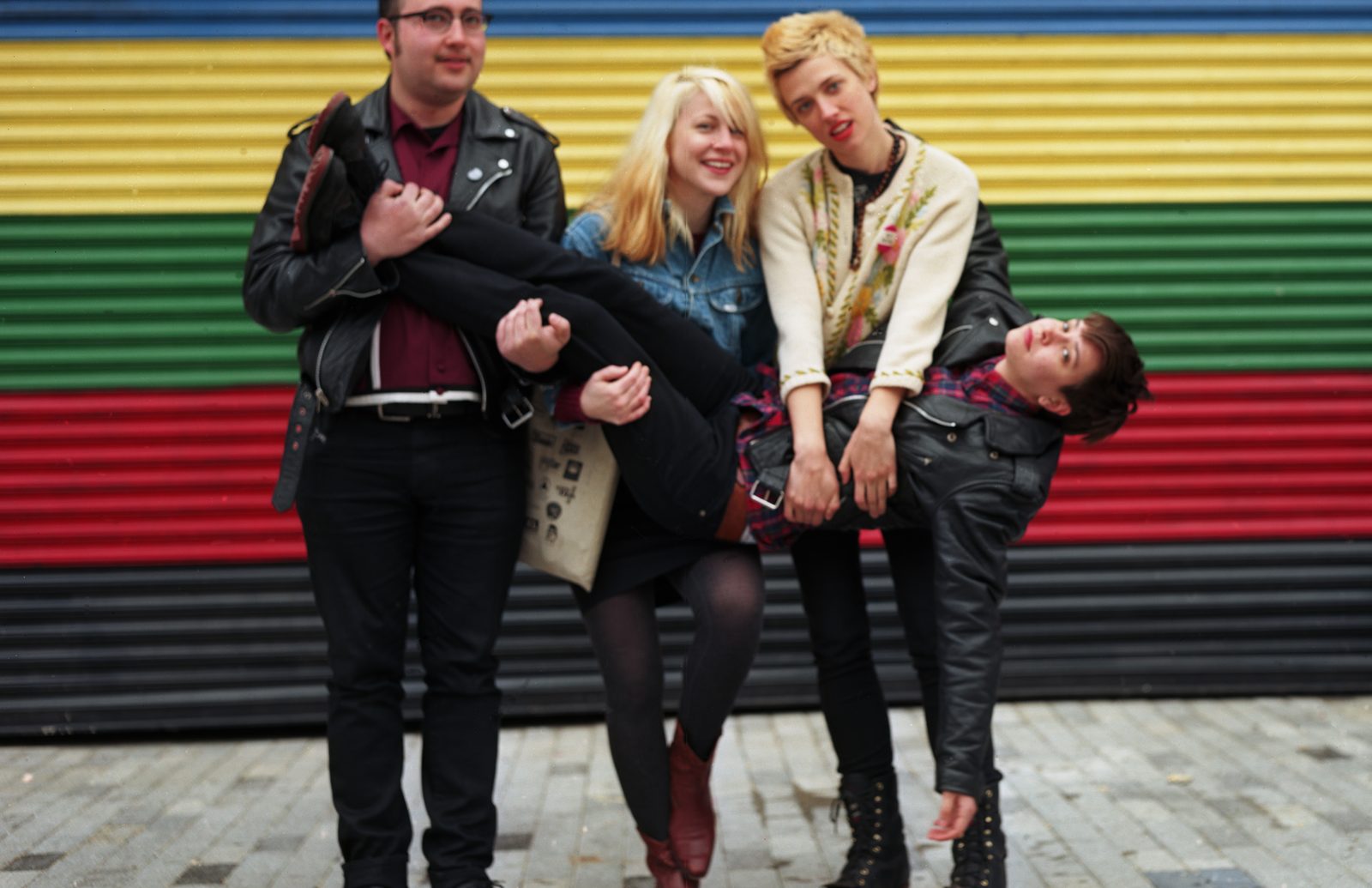 Priests clowning in front of a striped yellow, green, red and black shutter, three band members holding a fourth sideways in their arms