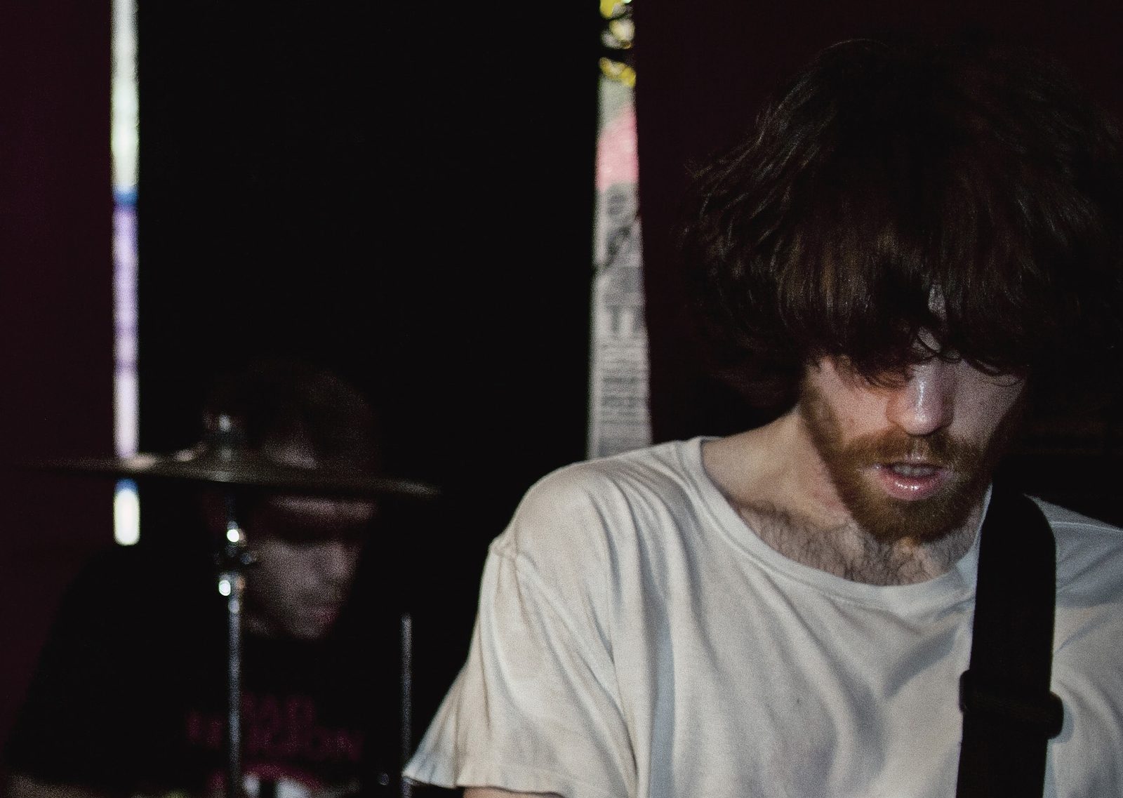 A bearded Boardroom Heroes guitarist with shaggy hair over his eyes in a white tee, the drummer behind him