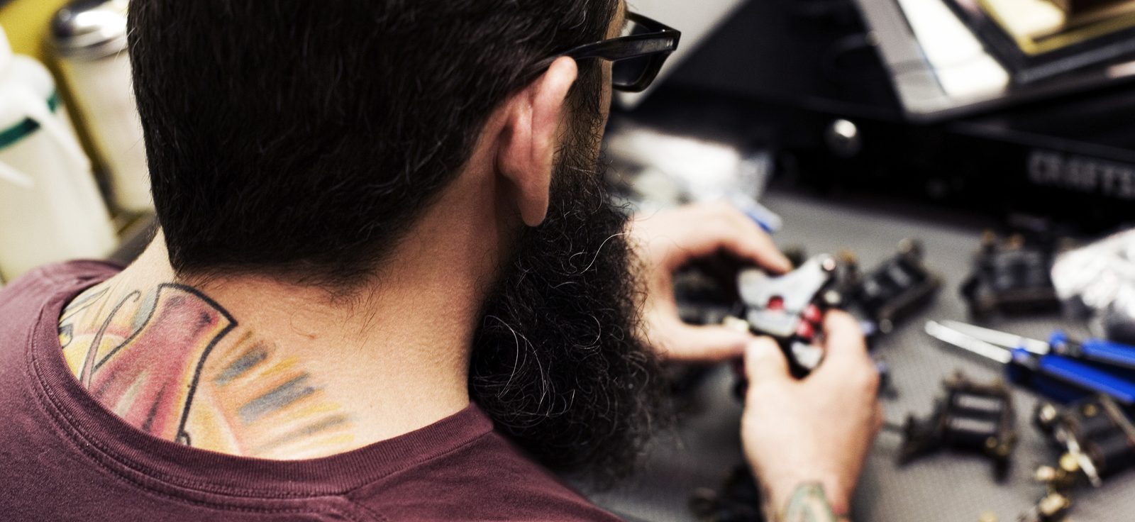 Bearded tattoo artist with a tattooed neck seen from behind, assembling a tattoo machine at his workstation