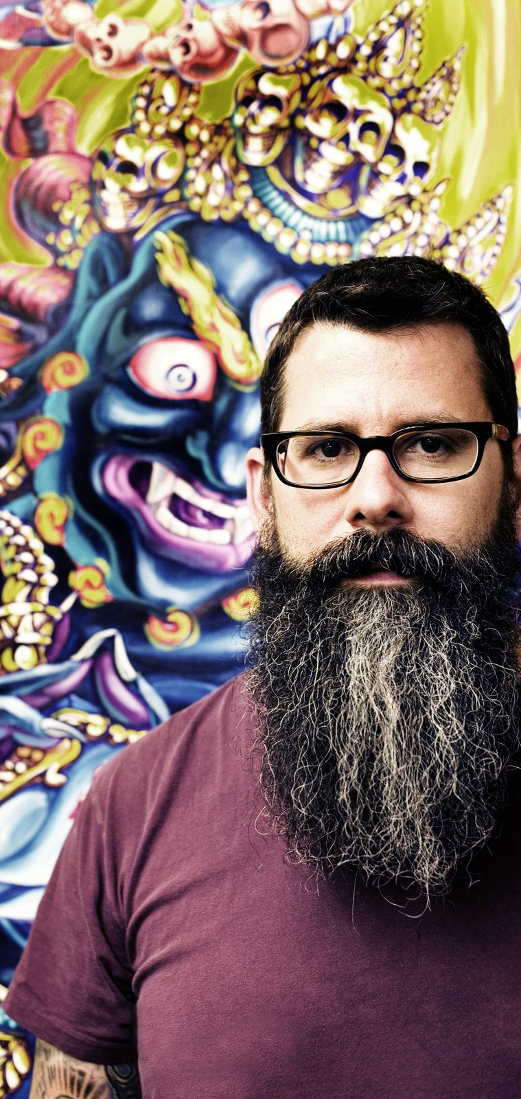 Bearded tattoo artist in glasses and a maroon tee in front of a colorful painted demon mask