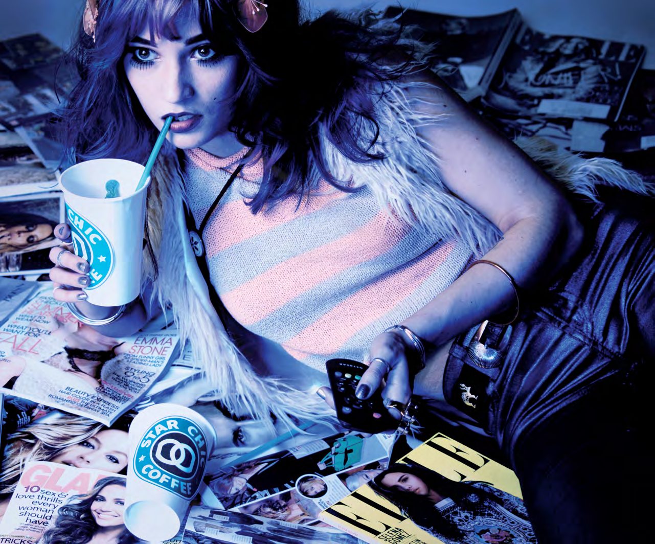 Model with dark hair and a pastel striped top sprawled across a pile of fashion magazines, sipping from a coffee cup in cool blue light