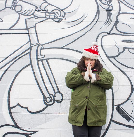 Woman in a green parka and a red knit hat presses her palms together in front of a black and white mural