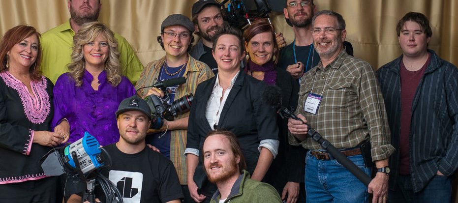 A large group of cast and crew pose together in front of a curtain, two women in purple at the front