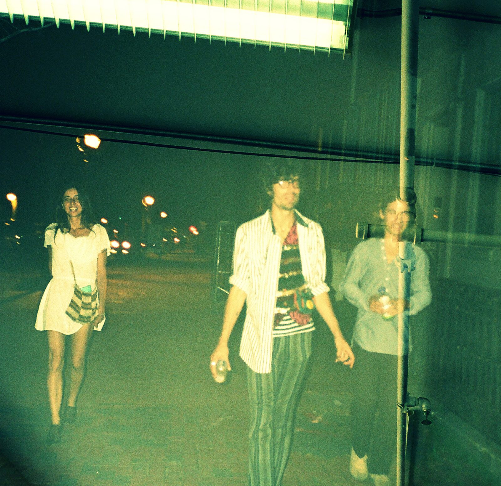 The three members of The Entrance Band walking down a night street under green fluorescent light, Guy Blakeslee in striped pants in the center