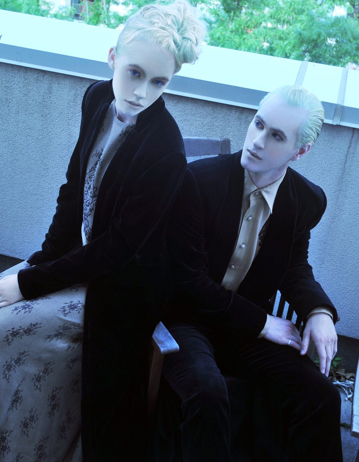 Two platinum-haired models in dark velvet tuxedo jackets and silk shirts seated on a terrace