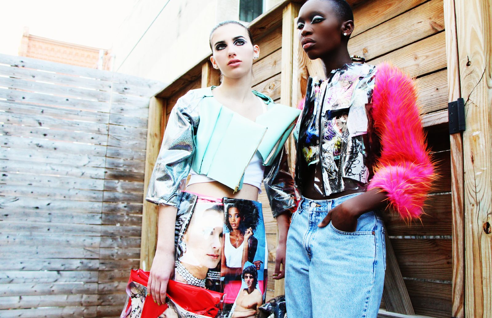 Two models against a wooden fence and gray boards, one in a mint cropped jacket and magazine collage skirt, the other in a collage jacket with hot pink faux fur sleeves and light wash jeans