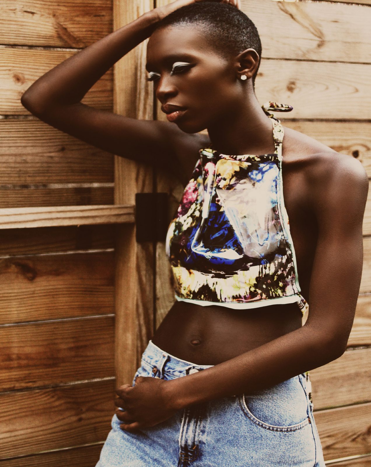Model with one arm raised behind her head in a painted print halter crop top and light wash jeans against wooden boards