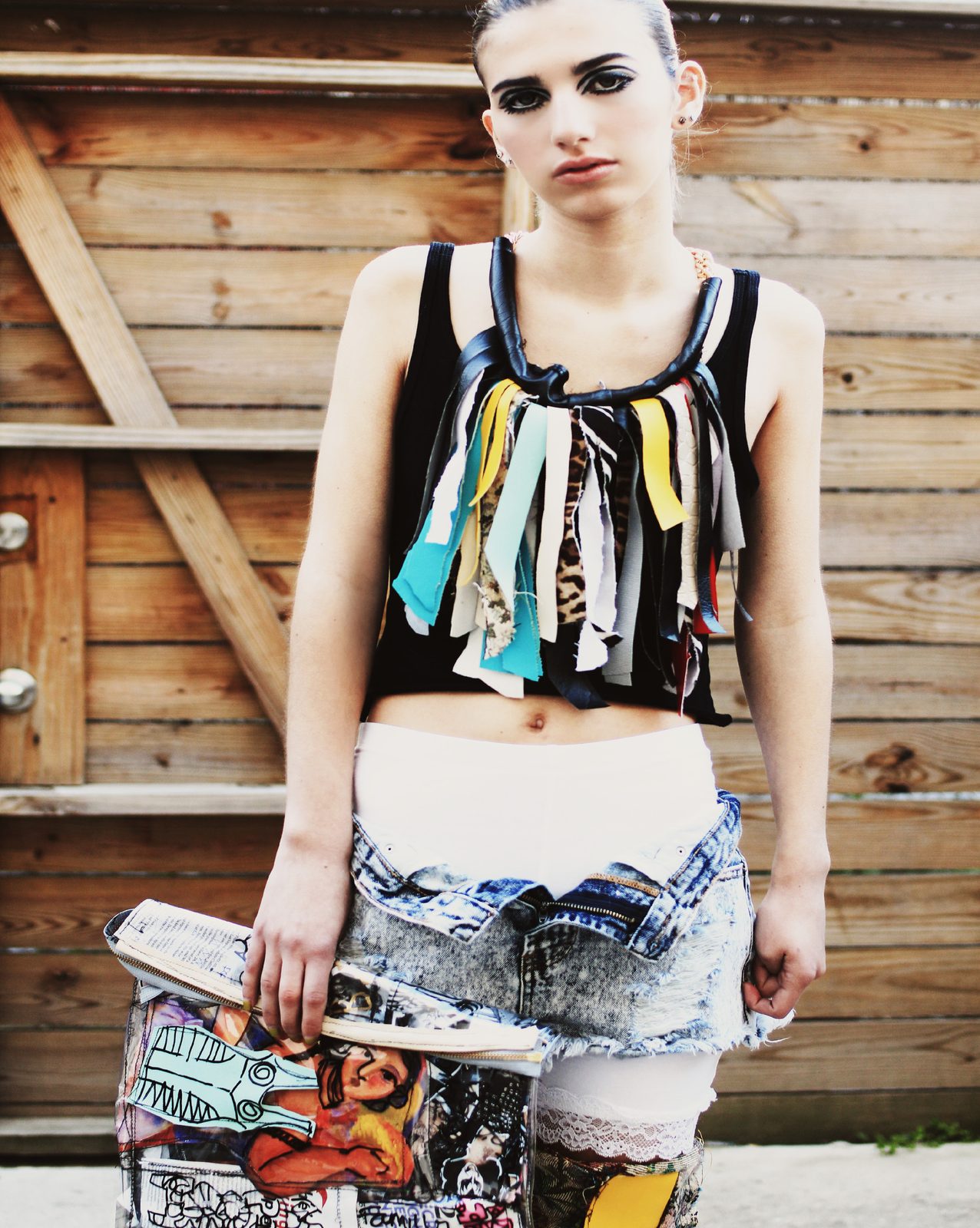 Model in a black tank with a fringed collage bib necklace, acid wash shorts and patchwork leggings, holding a comic print clutch