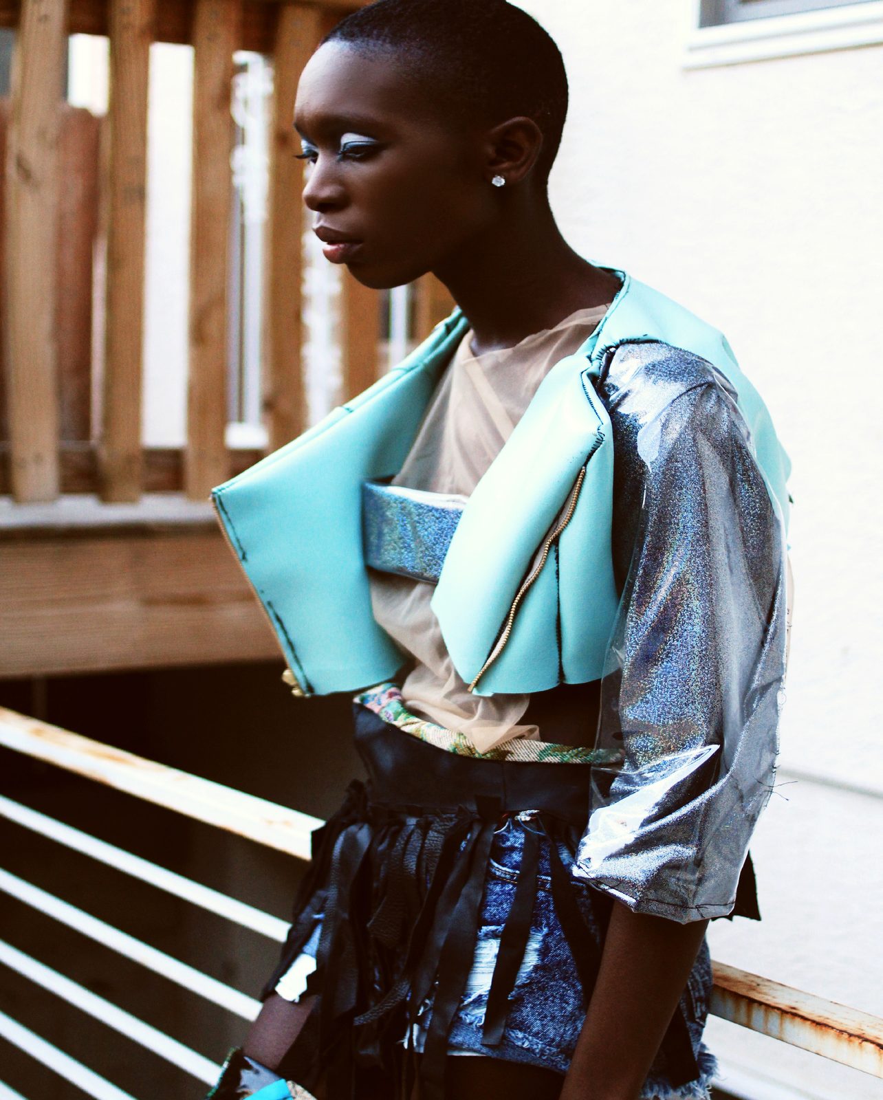 Model in a structured mint and metallic silver cropped jacket over a sheer top, standing by a wooden stair rail