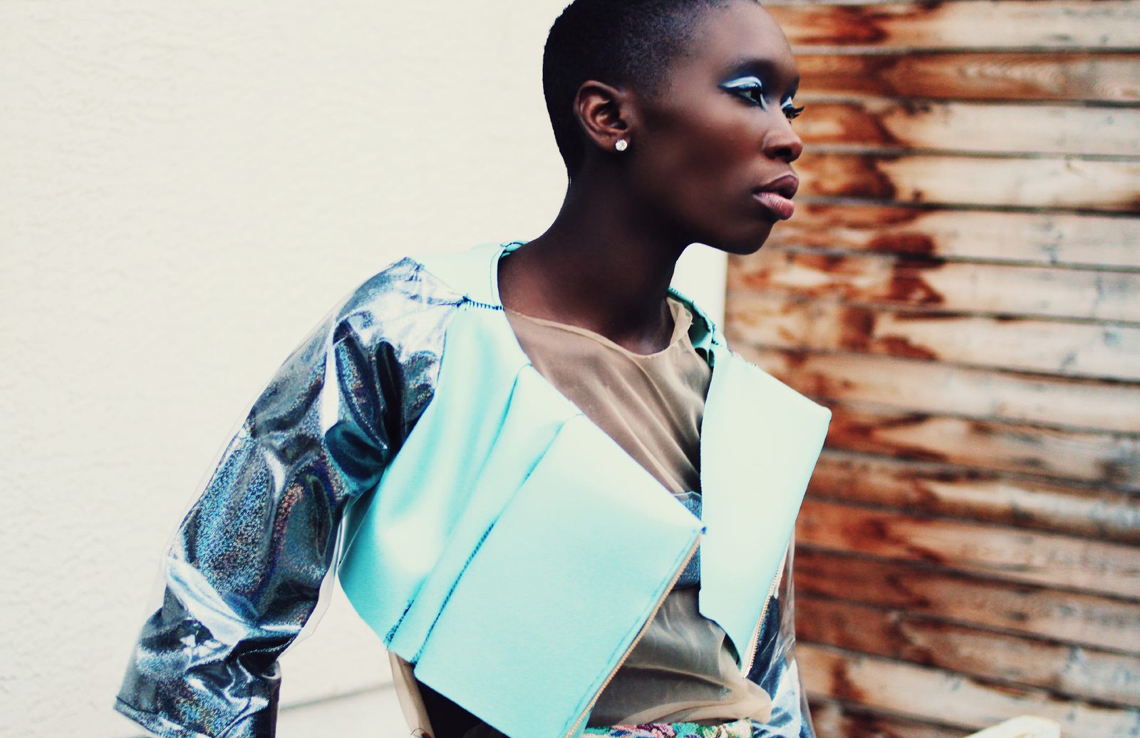 Wide shot of a model in a mint and silver metallic cropped jacket against a white wall and rusted wooden boards