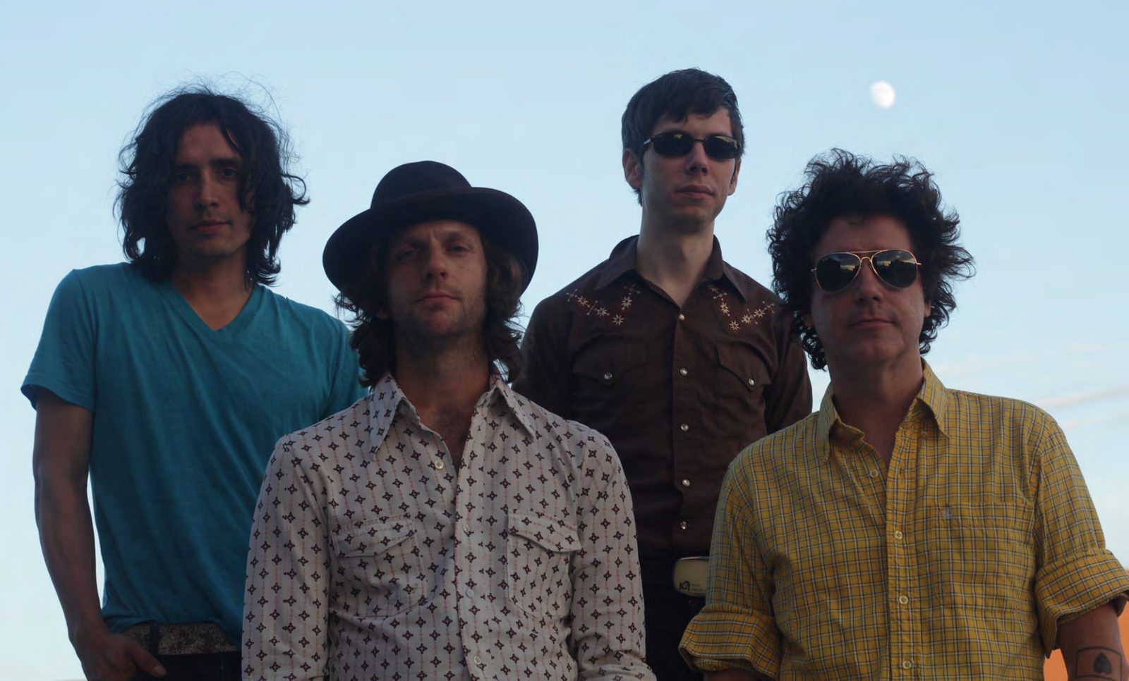 Four members of The Archive in vintage western shirts and sunglasses standing against a pale evening sky