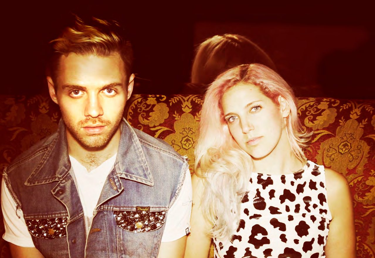Duo MS MR, a man in a denim vest and a platinum blonde woman in a leopard print top, seated against a dark red backdrop