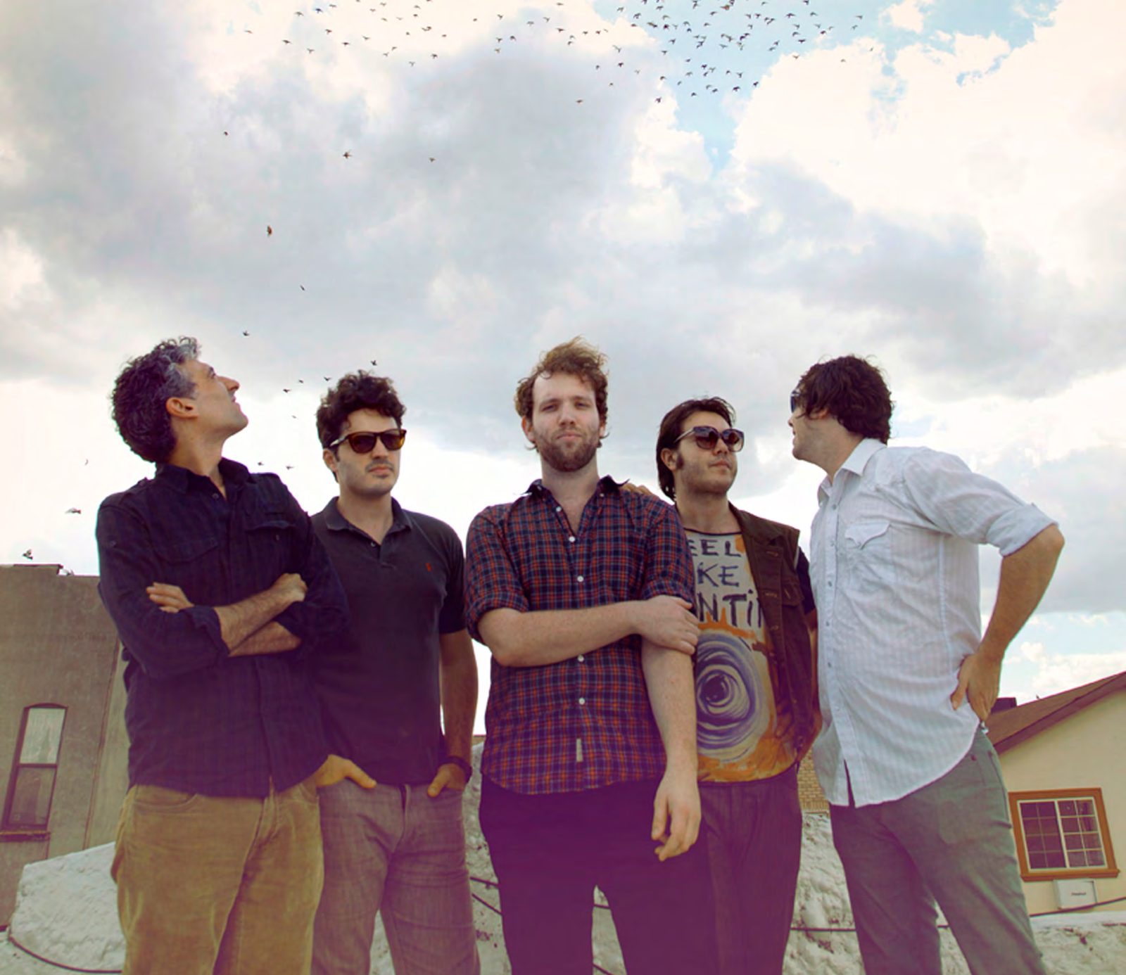 Five members of the band Caveman standing on a rooftop under a cloudy sky full of birds