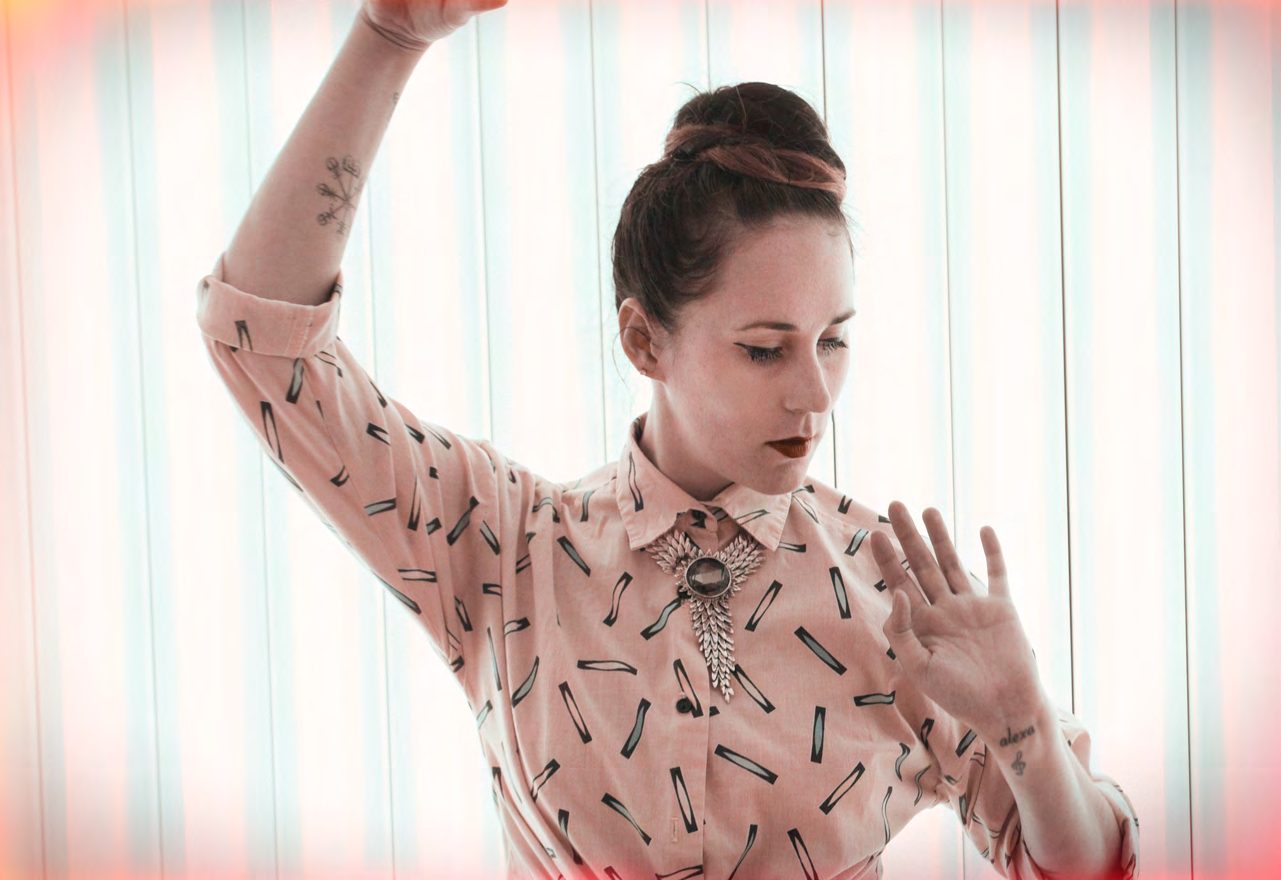 Johanna of Johanna and The Dusty Floor in a patterned blouse and bolo tie, one arm raised, against a striped pastel backdrop