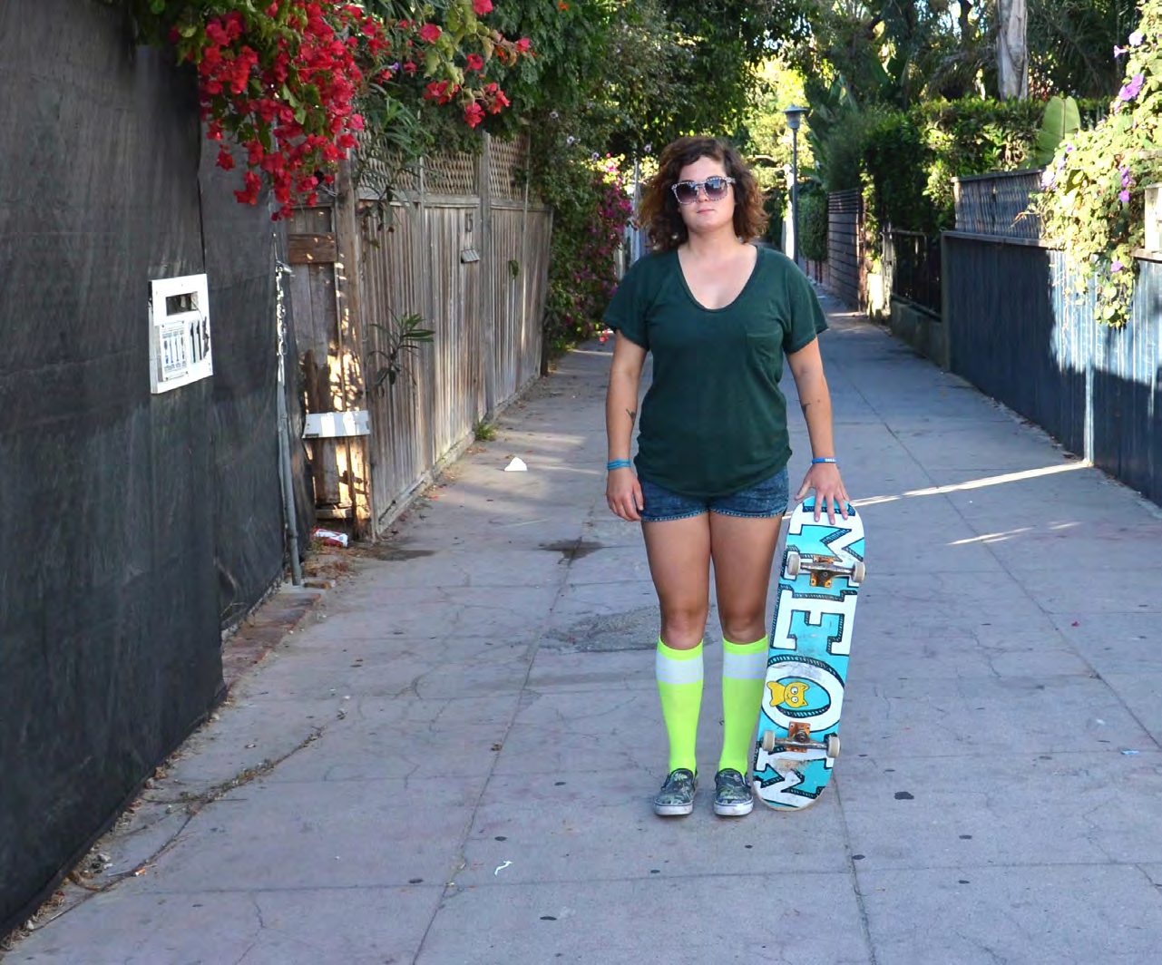 Skateboarder Amy Caron in sunglasses, a green tee, denim shorts and neon knee socks holding a MEOW skateboard in a leafy alley