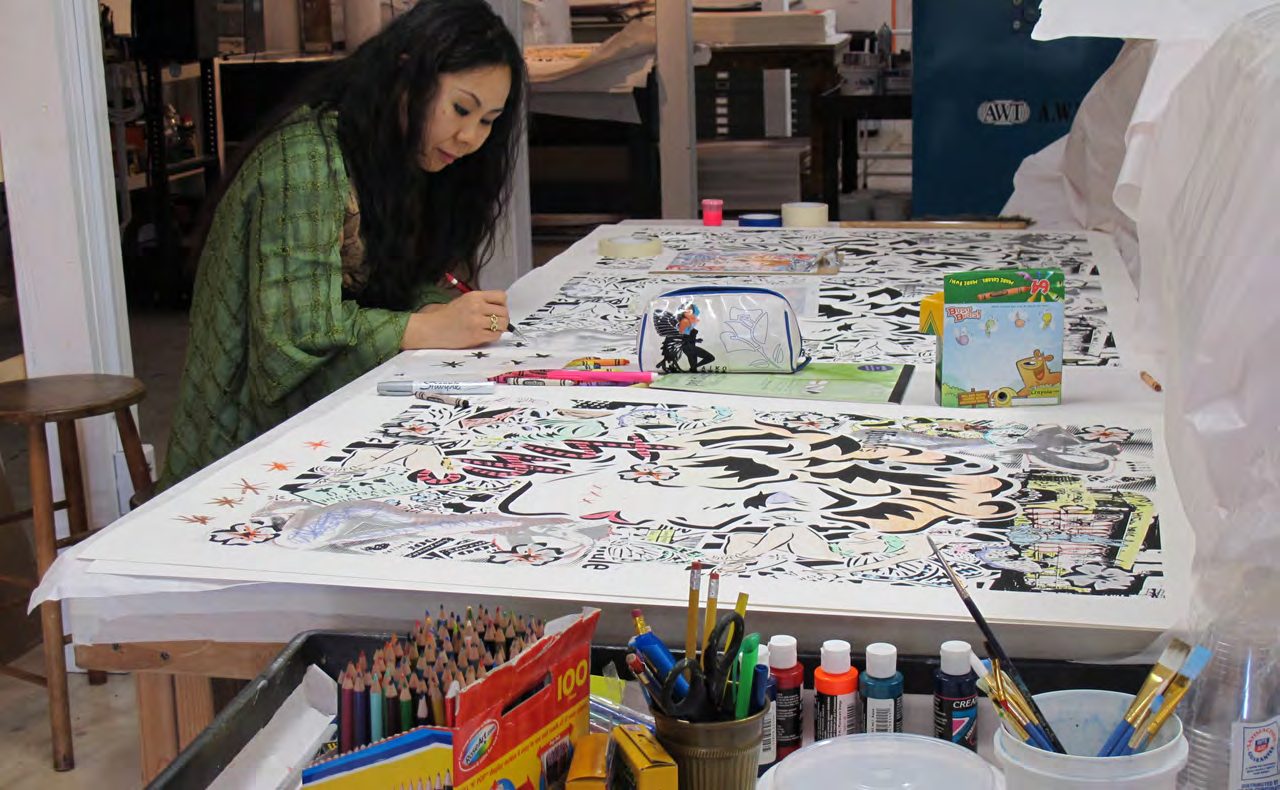 Artist Lady AIKO working at a long table covered in brightly colored paintings in her studio