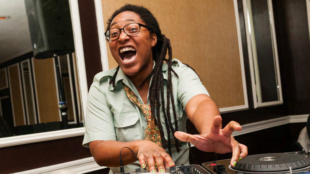 DJ Ebony Dumas, aka Natty Boom, laughing behind turntables in glasses and a khaki shirt
