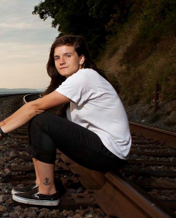 Skateboarder Kristin Ebeling in a white tee and black jeans sitting on railroad tracks at dusk