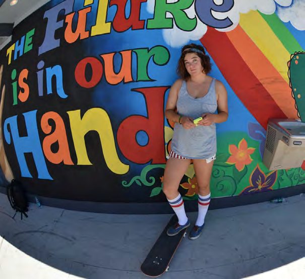 Skateboarder Amy Caron in a grey tank and striped tube socks standing with her board in front of a rainbow mural reading The Future is in our Hands