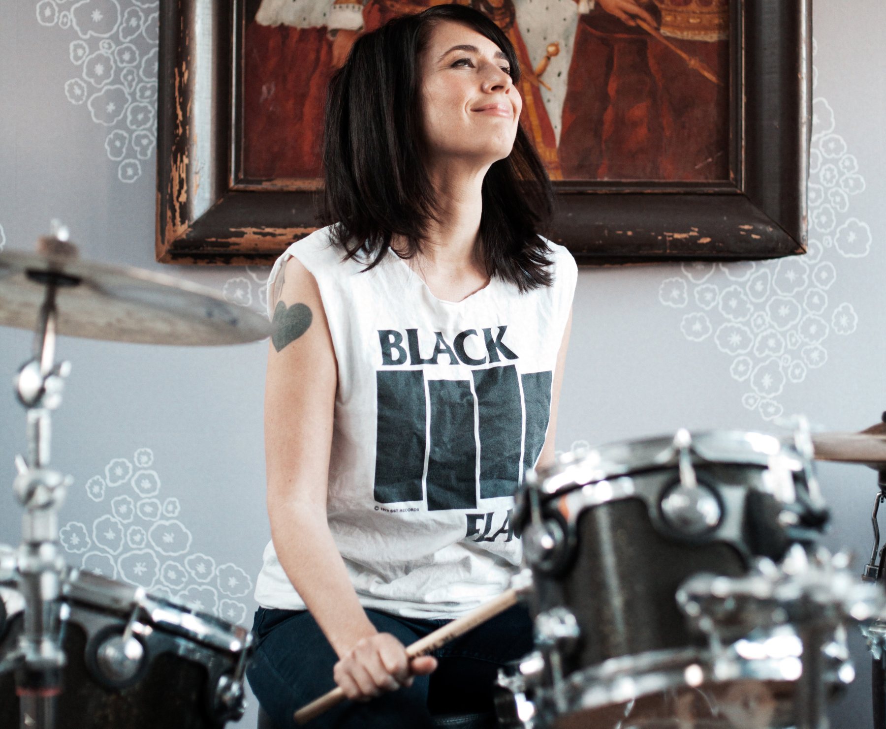 Kathleen Hanna smiling behind a drum kit in a Black Flag tank top, a framed painting on the wall behind her