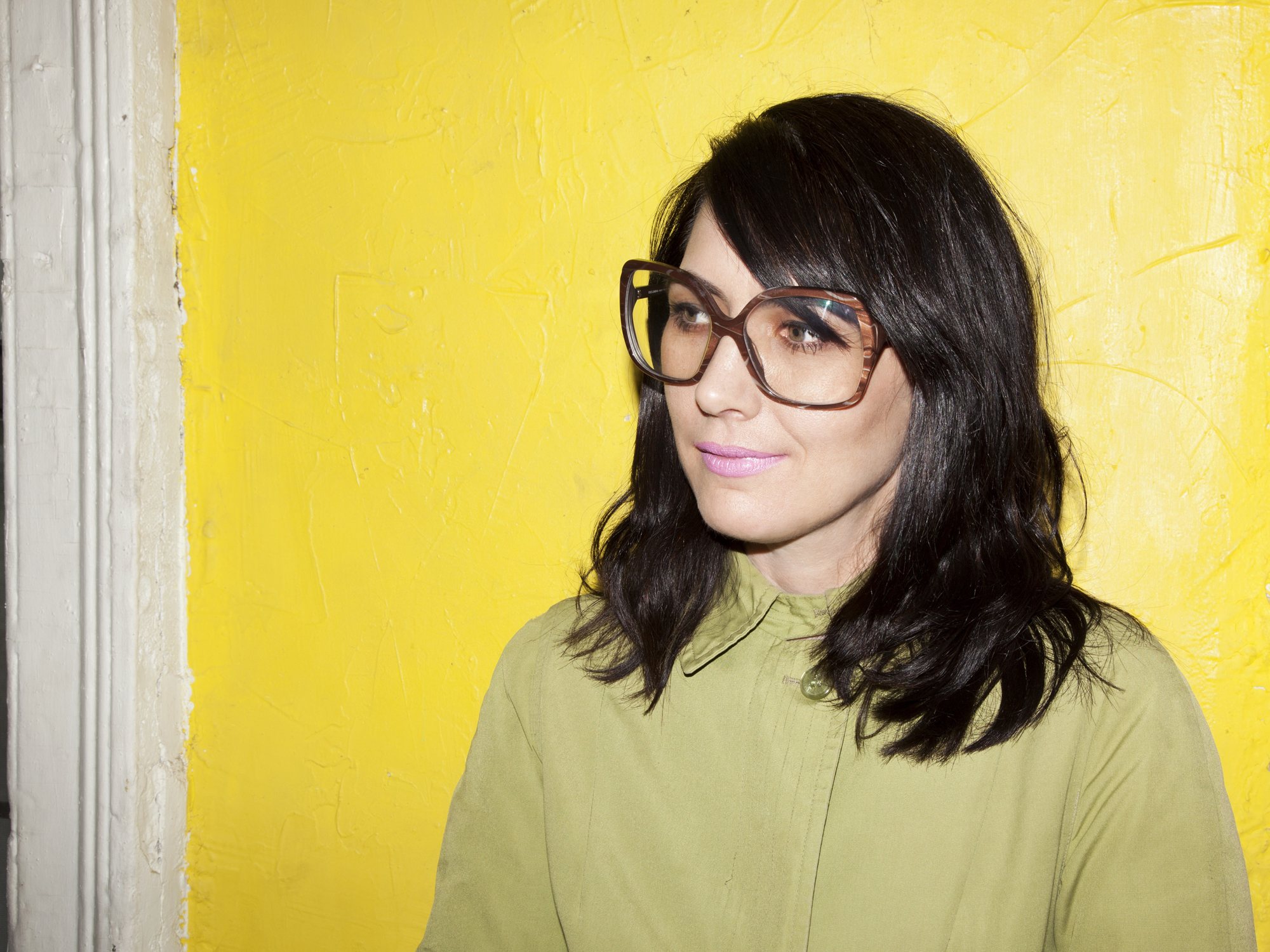 Kathleen Hanna in large tortoiseshell glasses and an olive shirt, against a bright yellow wall