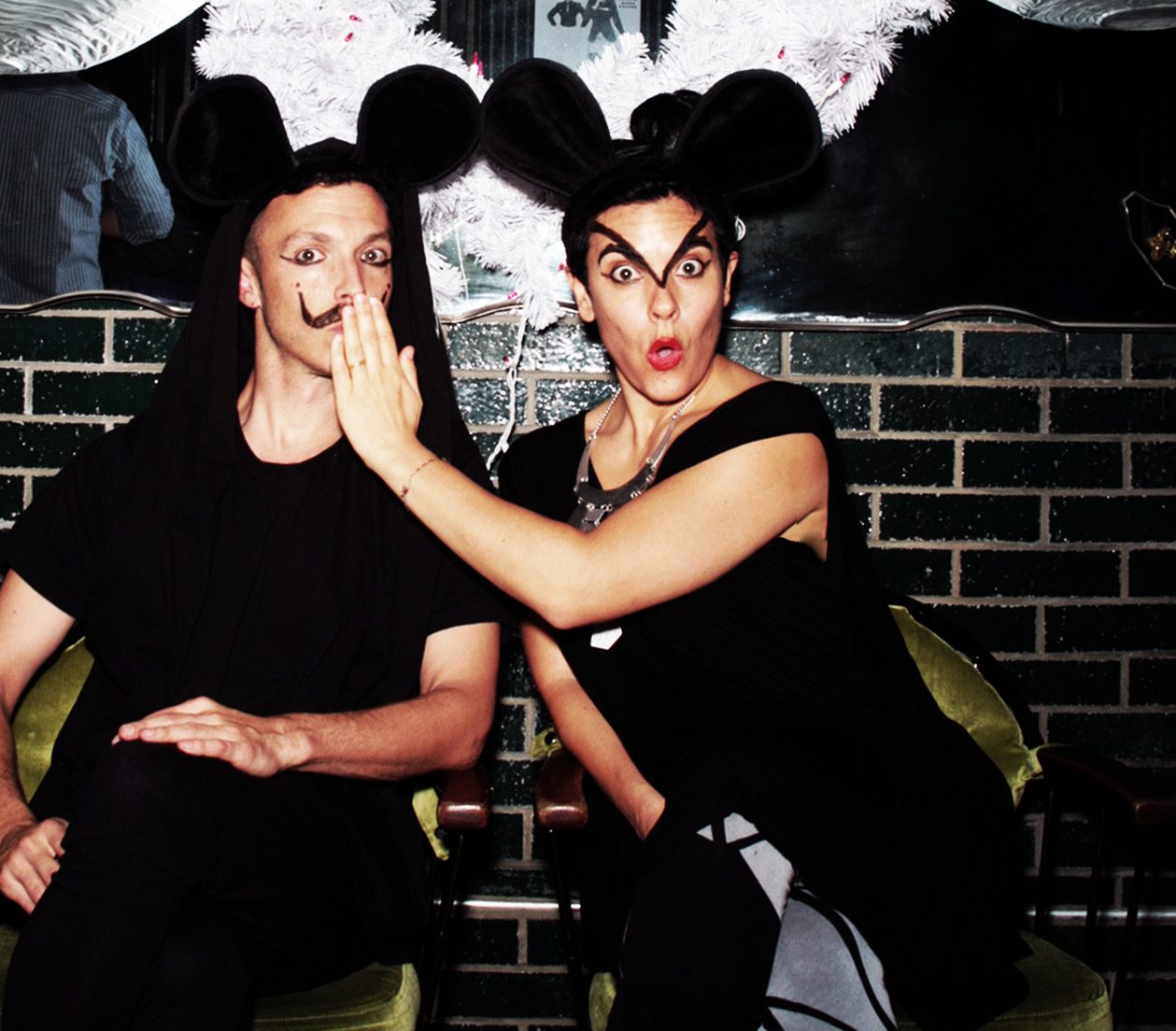Rick Gradone and Jen DM in black outfits and mouse ears, she covers his drawn-on mustache with her hand and makes a shocked face
