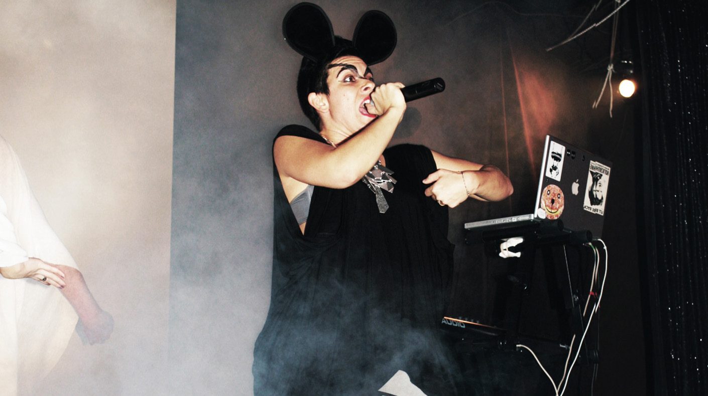 Jen DM in black mouse ears and a sleeveless black top rapping into a microphone on a smoky stage beside a laptop