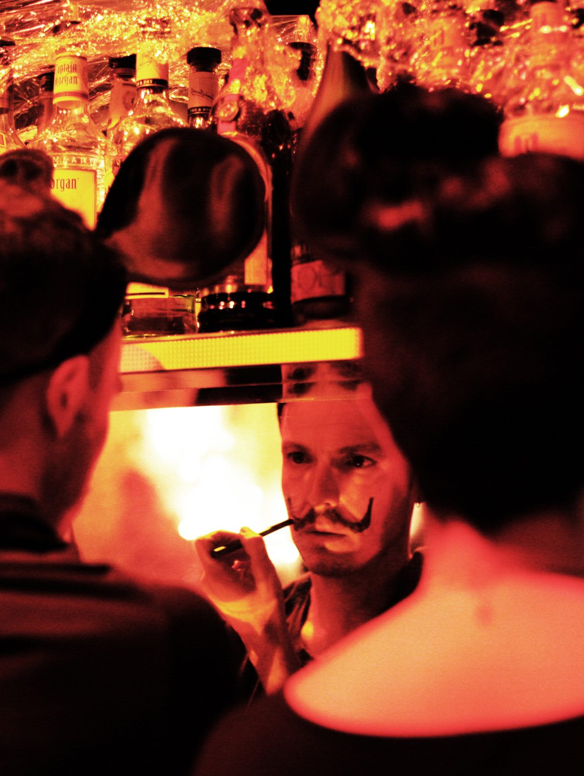 Rick Gradone with a drawn-on mustache, lit orange in a crowded bar