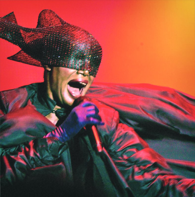 Grace Jones mid-song on stage in a sparkling black fish-shaped headpiece and billowing red cape against an orange backdrop