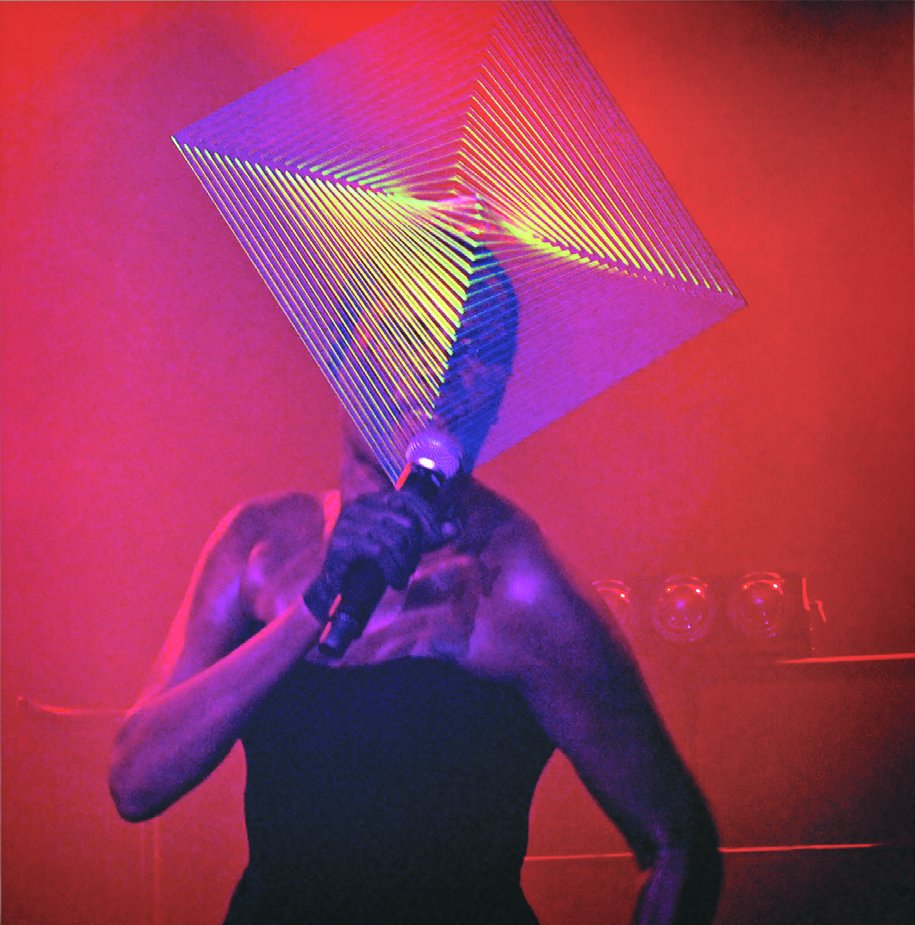Grace Jones singing into a microphone on stage under red light, her face hidden behind a huge geometric string-art headpiece