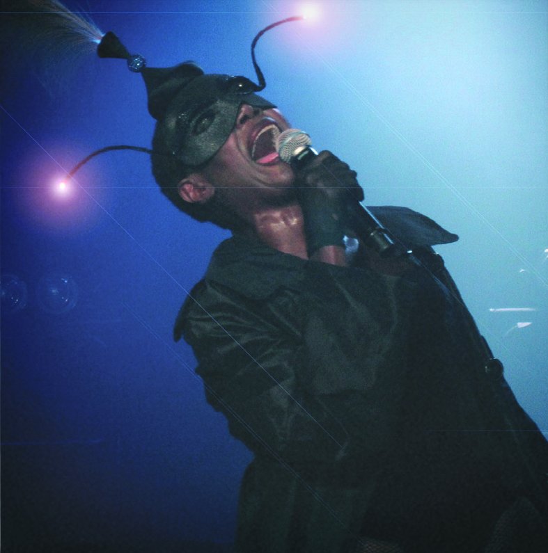 Grace Jones in a black leather coat and antenna headpiece, head thrown back, singing into a microphone under blue stage light