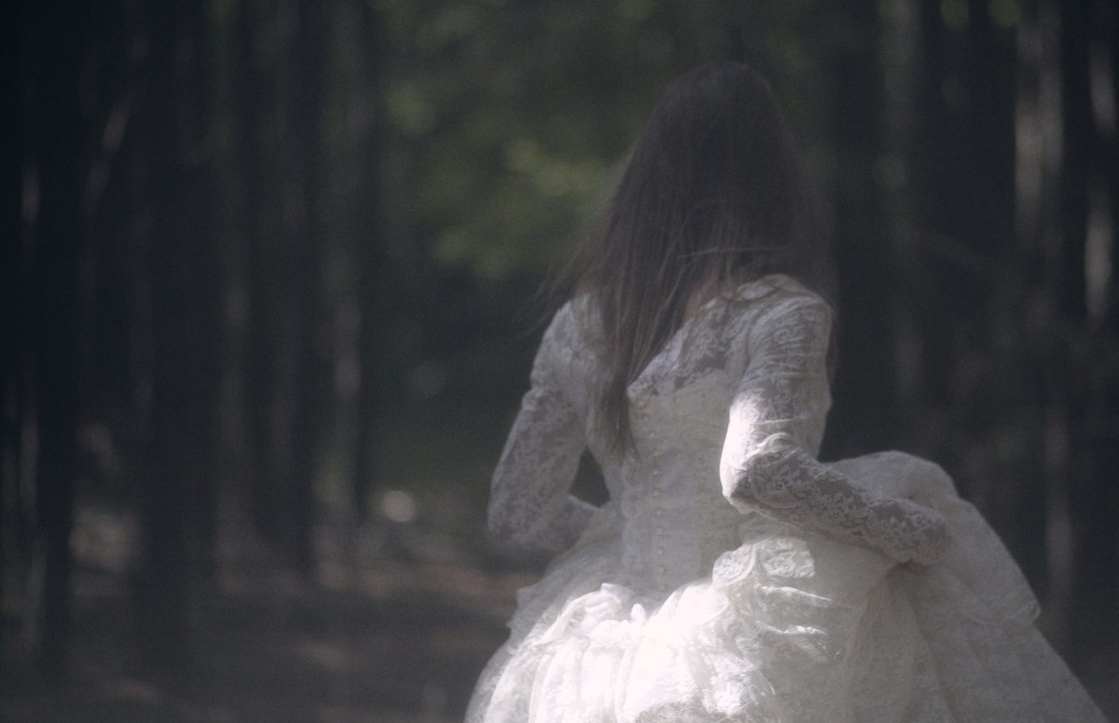 Model in a long-sleeved white lace gown runs away from the camera down a dim forest path, hair streaming
