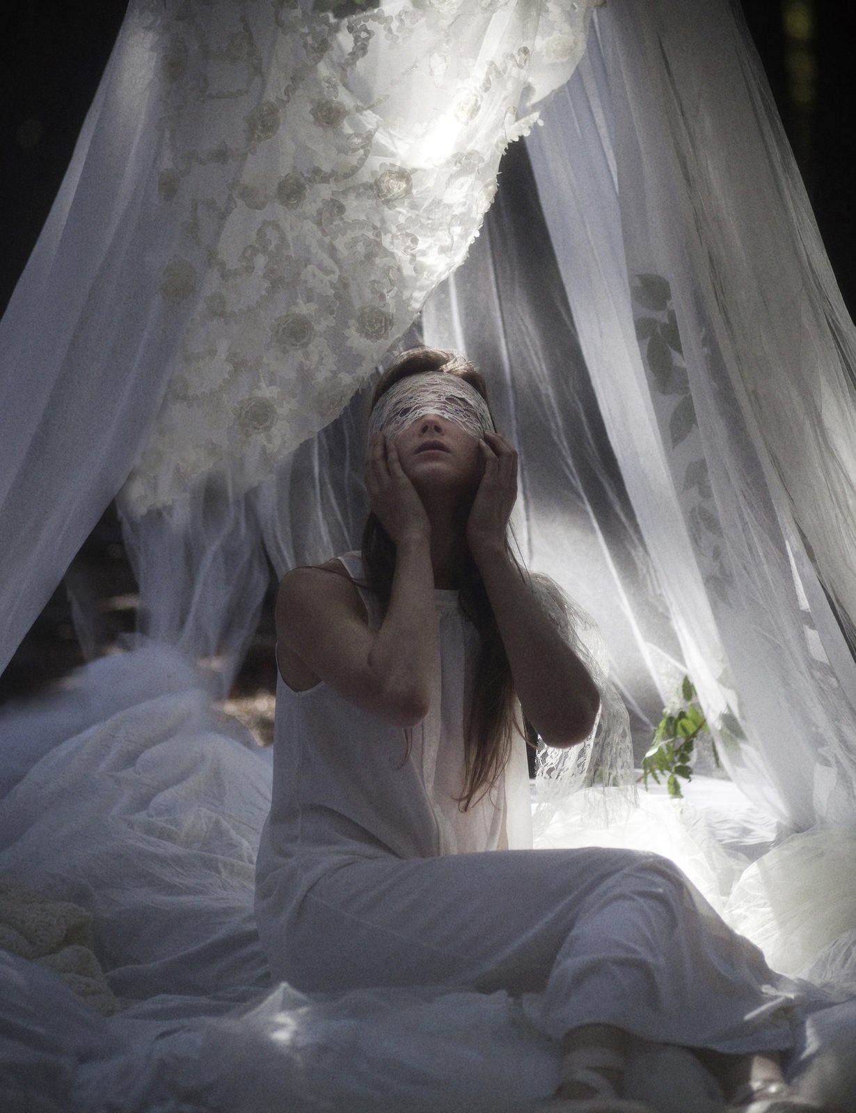 Model with eyes closed and hands to her face sits under a white canopy draped with flowers