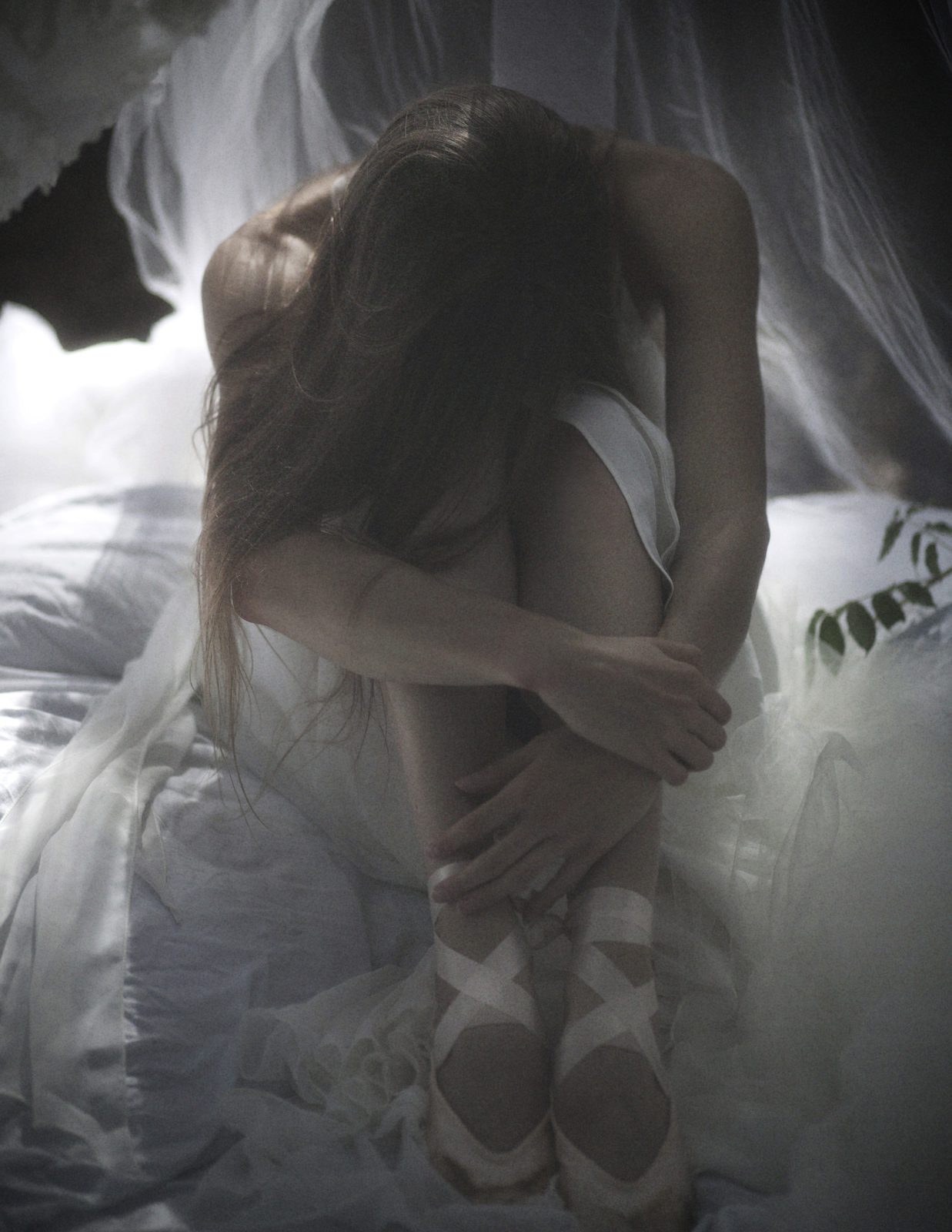 Model curled up with her head on her knees, wearing satin ballet shoes, in white tulle