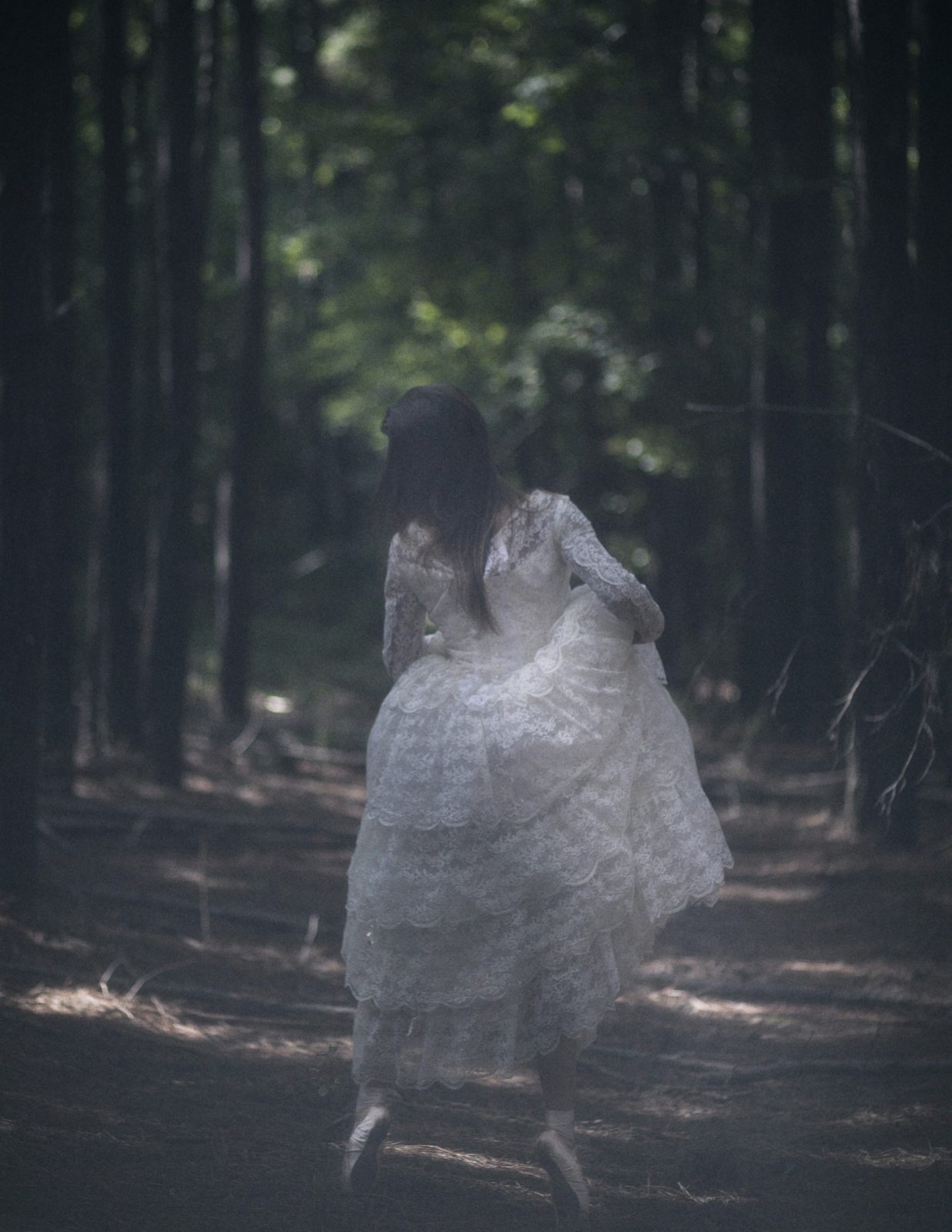 Model in a tiered white lace dress walks away down a forest path