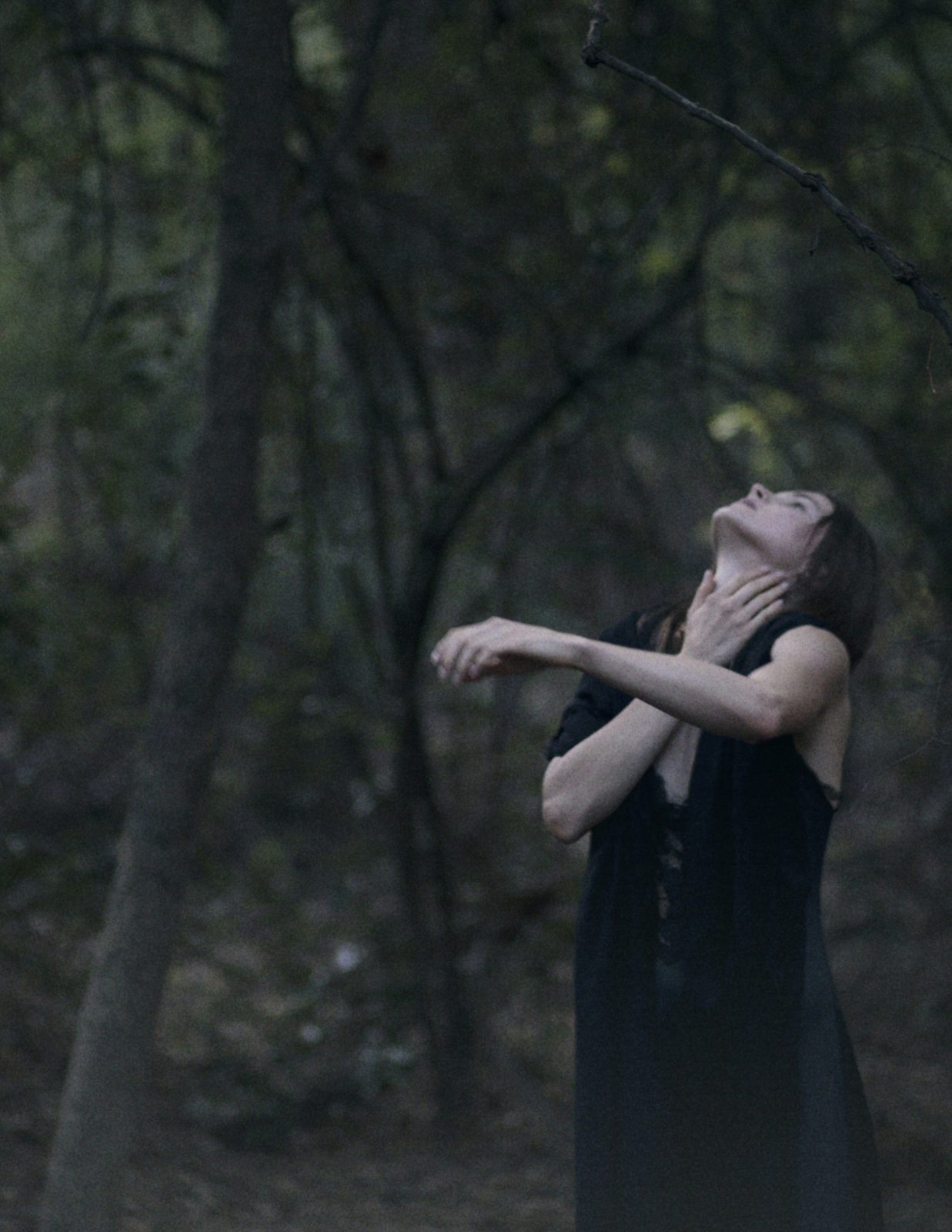 Model in a black dress tilts her head back and clutches her throat among bare trees