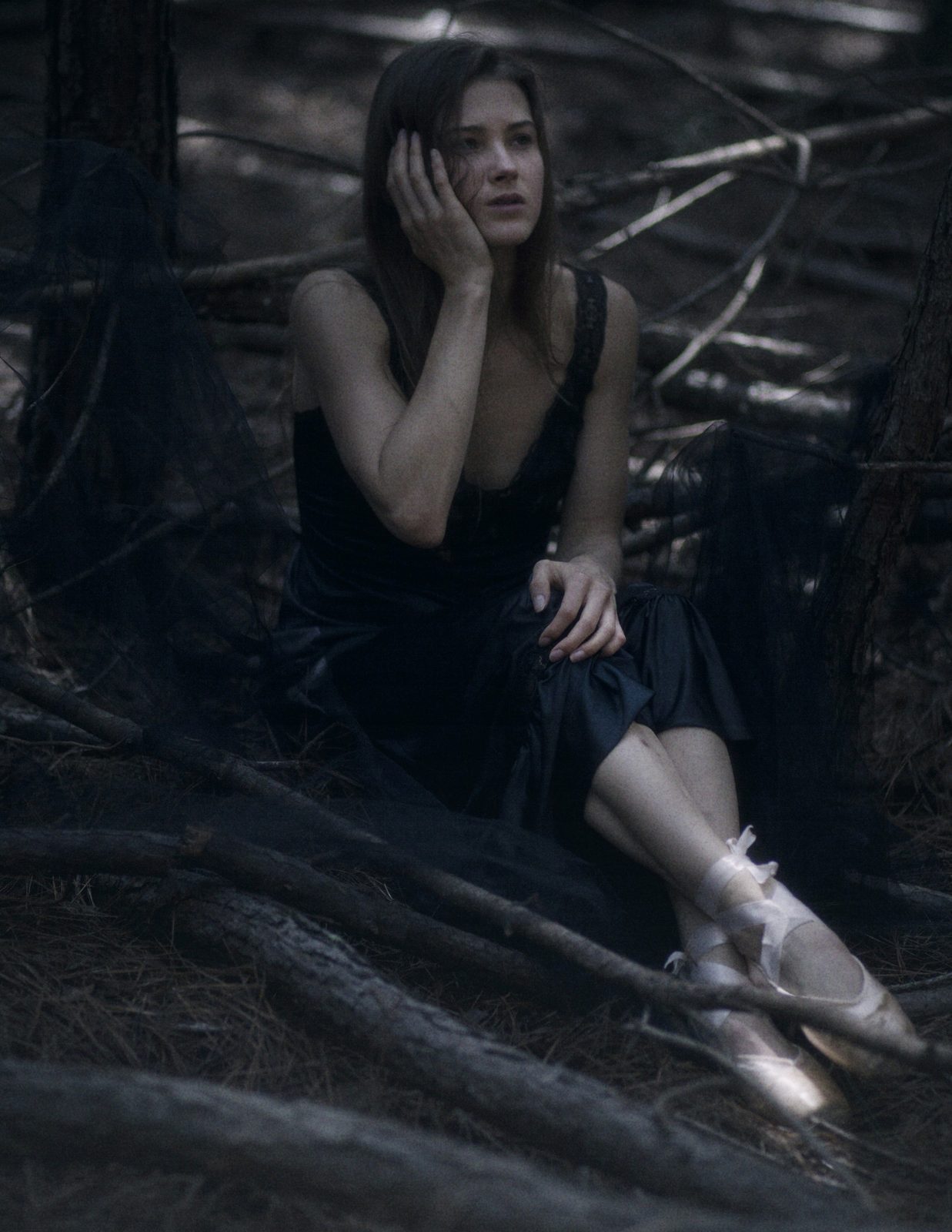 Model in a black dress and ballet shoes sits among fallen branches, hand to her cheek