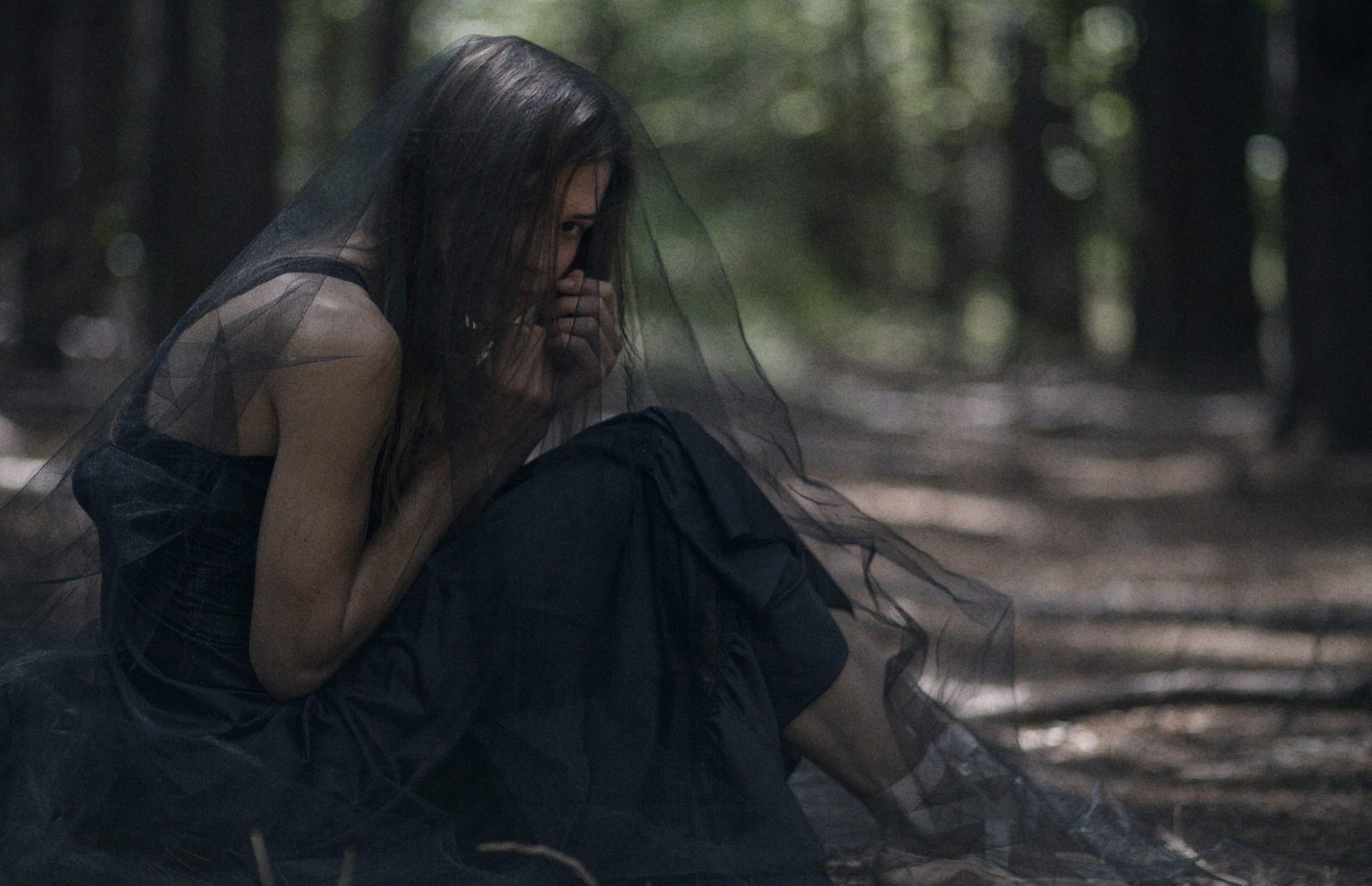 Model in a black dress and black tulle veil crouches on a sunlit forest floor, hands at her mouth