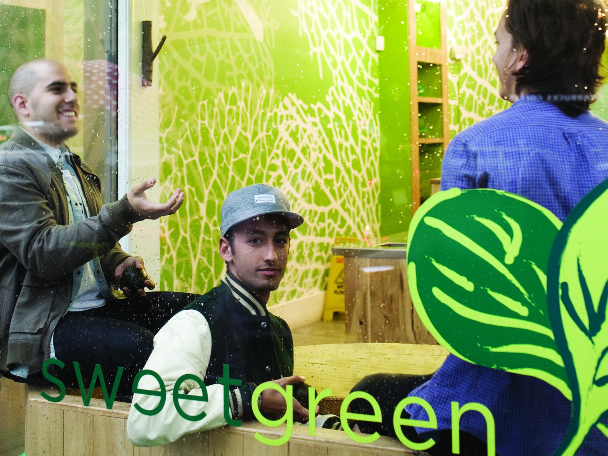 Sweetgreen founders Nicolas Jammet, Jonathan Neman and Nathaniel Ru laughing and talking inside a bright green restaurant with the Sweetgreen logo on the glass