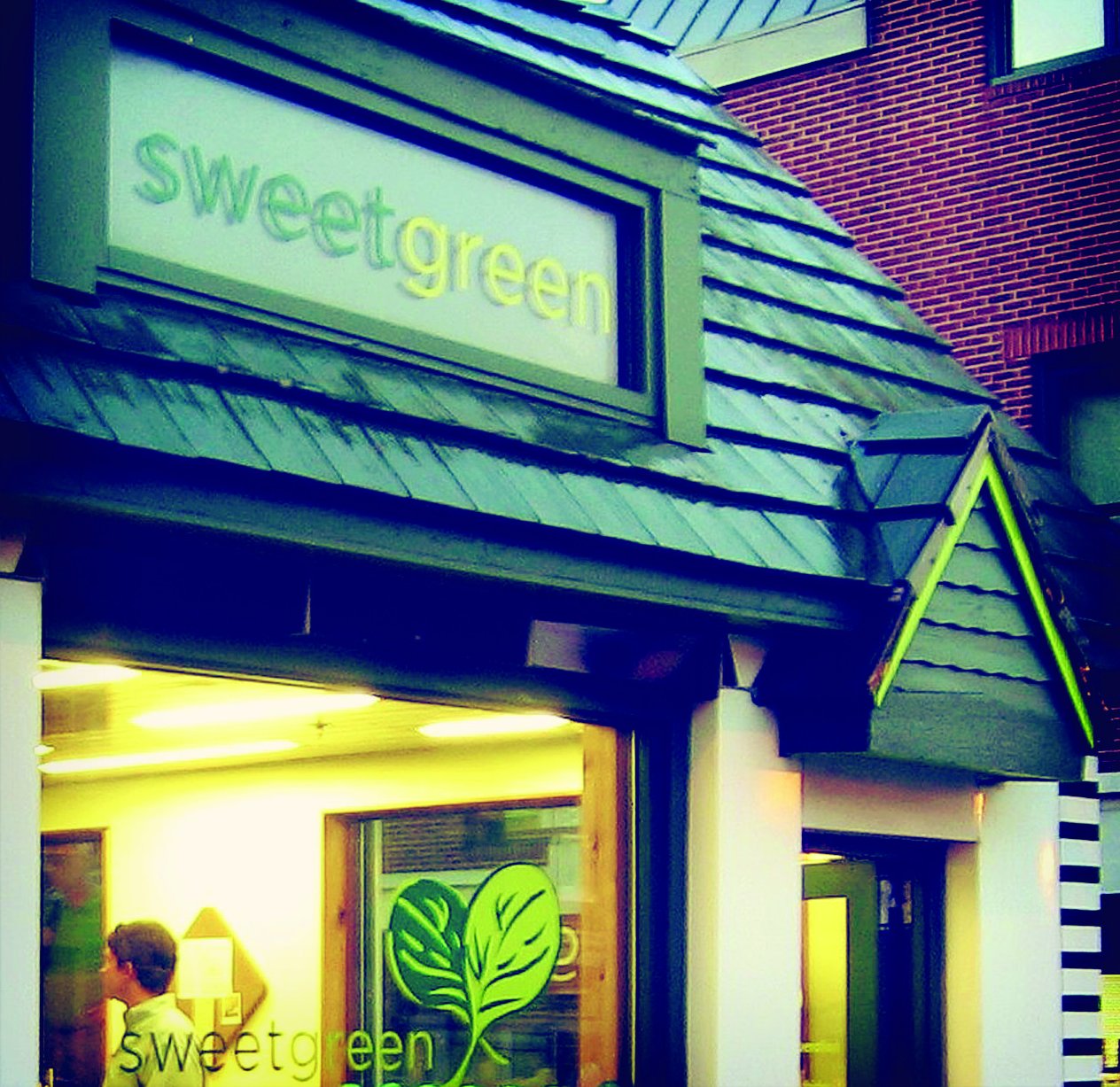 Sweetgreen storefront with a green shingled awning, glowing windows and the leaf logo on the glass