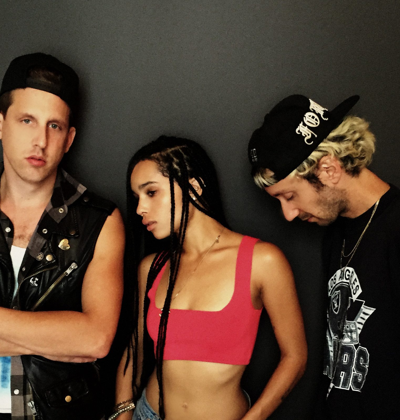 Zoë Kravitz in a red crop top with long braids standing between her two LolaWolf bandmates, one in a black cap and sleeveless studded vest, the other in a black cap and black graphic tee, against a dark gray wall