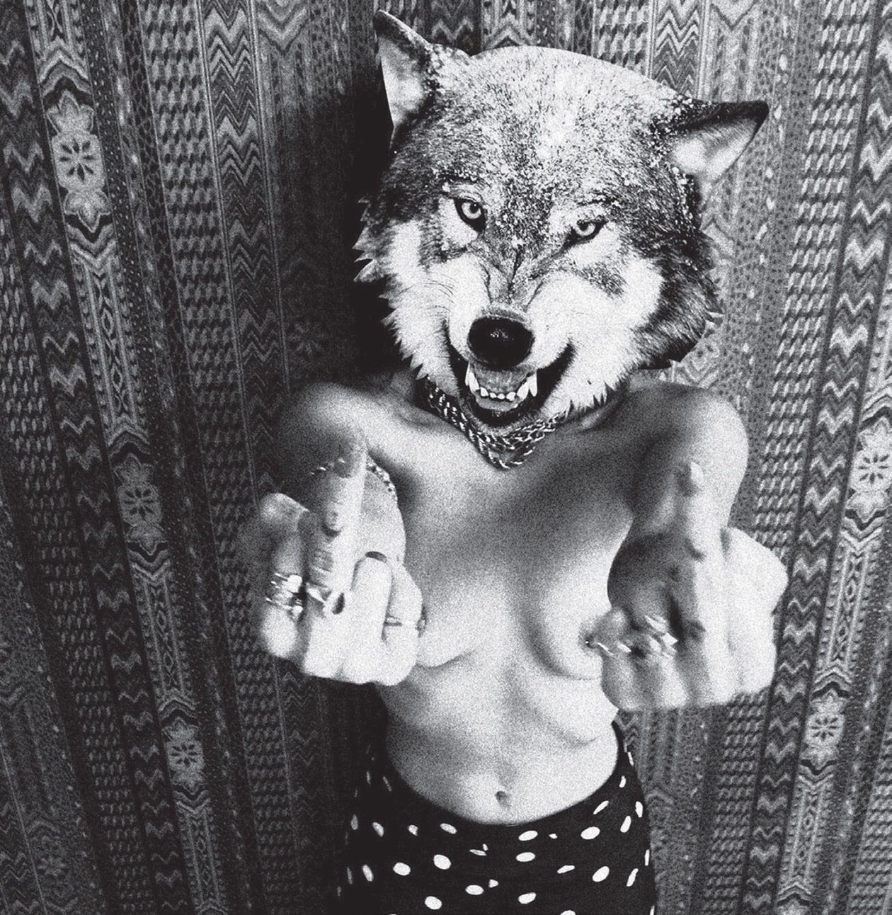 Black and white photo of a topless figure in a snarling wolf head mask and spiked collar, wearing rings and polka dot bottoms, flipping off the camera with both hands in front of a patterned textile wall