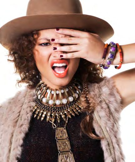 Model laughing with one hand over her eye, wearing a brown fedora, a stack of bead and pearl necklaces and Peruvian bracelets